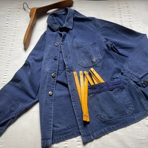 May include: A dark blue long-sleeve jacket with a collar, front button closure, and multiple patch pockets. The jacket features a worn texture and is displayed with a wooden hanger and a yellow folding ruler measuring approximately 7.8 inches.