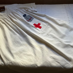 Può includere: Camice medico in cotone bianco con una croce rossa e una bottiglia blu e bianca con la scritta "To Self" stampata su di essa.