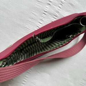 May include: A pink crossbody bag with a zippered main compartment. The interior lining is green and white striped. The bag has a matching pink strap and silver-toned hardware. The bag is open, revealing the interior pockets.
