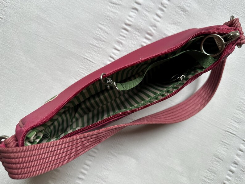 May include: A pink crossbody bag with a zippered main compartment. The interior lining is green and white striped. The bag has a matching pink strap and silver-toned hardware. The bag is open, revealing the interior pockets.