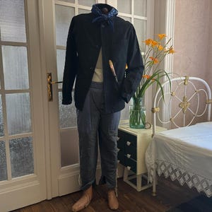 May include: A black jacket, light-colored shirt, and blue pants are displayed on a mannequin. A blue patterned scarf is around the neck. A paintbrush is in the pocket. The mannequin is in front of a white door and a bed.