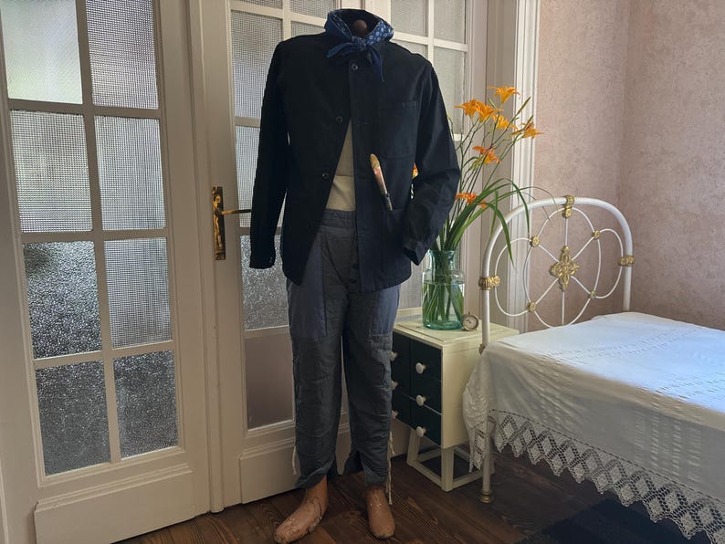 May include: A black jacket, light-colored shirt, and blue pants are displayed on a mannequin. A blue patterned scarf is around the neck. A paintbrush is in the pocket. The mannequin is in front of a white door and a bed.