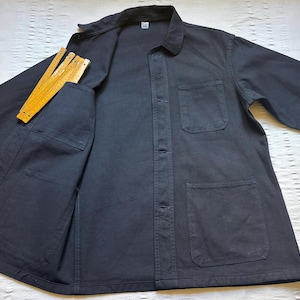 May include: A navy blue work jacket with a collared neckline, button-up front, and multiple pockets. A yellow folding ruler is in the chest pocket. The jacket is made of a durable fabric.