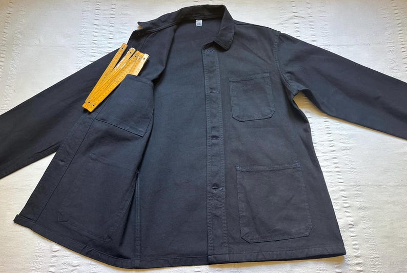 May include: A navy blue work jacket with a collared neckline, button-up front, and multiple pockets. A yellow folding ruler is in the chest pocket. The jacket is made of a durable fabric.