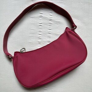 May include: A magenta shoulder bag with a curved shape and a matching strap. The bag has a zipper closure and a textured surface. The strap is attached with silver-colored metal hardware.