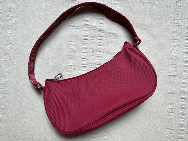 May include: A magenta shoulder bag with a curved shape and a matching strap. The bag has a zipper closure and a textured surface. The strap is attached with silver-colored metal hardware.