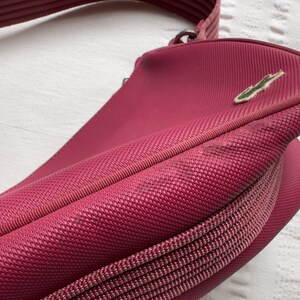 May include: A close-up of a red-pink crossbody bag with a textured fabric. The bag has a zipper closure and a shoulder strap. A green crocodile logo is visible on the front.