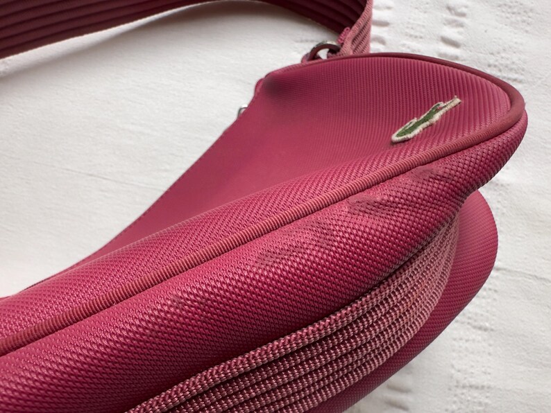 May include: A close-up of a red-pink crossbody bag with a textured fabric. The bag has a zipper closure and a shoulder strap. A green crocodile logo is visible on the front.