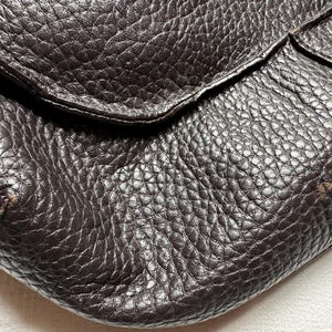 May include: Close-up of a dark brown leather bag with a textured surface. The bag has a flap closure and a slightly worn appearance, with visible grain and subtle imperfections.