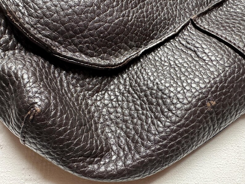 May include: Close-up of a dark brown leather bag with a textured surface. The bag has a flap closure and a slightly worn appearance, with visible grain and subtle imperfections.