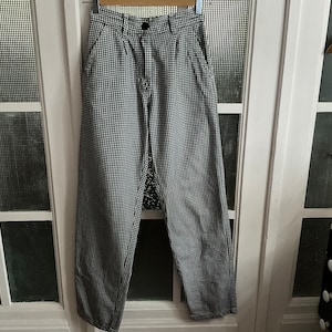 May include: Black and white checkered pants hanging on a wooden hanger. The pants have a button closure and pockets. The fabric has a small, repeating pattern. The pants are hanging in front of a white door with glass panes.