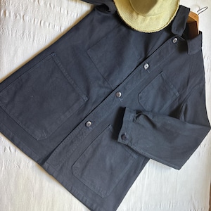 May include: A navy blue work jacket with four front pockets and button closures. A light tan straw hat rests on the jacket. The jacket is made of a sturdy fabric and is laid out on a white surface.