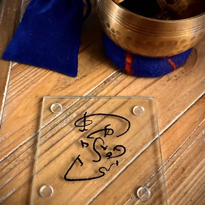 May include: A glass coaster with a black ink drawing of a symbol on it. The coaster has four clear rubber feet on the bottom. The coaster is sitting on a wooden surface.