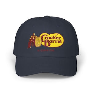 Op de afbeelding: Marineblauwe baseballcap met het Cracker Barrel-logo. Het logo heeft een geel en rood ontwerp met de woorden "Cracker Barrel" en een figuur die tegen een vat leunt. De pet heeft een gebogen klep.
