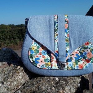 以下が含まれることがあります： 青いデニムのクロスボディバッグ。花柄の生地の裏地と銀色のバックル留め具が付いています。