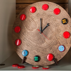 Könnte beinhalten: Eine Holzuhr mit einem Zahnrad-Design und Flaschenverschlüssen als Ziffern. Die Uhrzeiger sind schwarz und rot. Die Uhr steht auf einem weißen Regal mit verstreuten Flaschenverschlüssen. Das Zifferblatt der Uhr zeigt "WHY NOT" und "CAPS&BOARD.COM".
