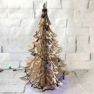 May include: A three-dimensional laser-cut wooden Christmas tree with intricate scrollwork patterns throughout its branches. The structure is wrapped in a string of small, multi-colored LED lights with a black cord, creating a decorative display.