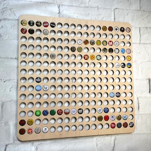 May include: A wooden bottle cap display board with many circular holes, each filled with a colourful bottle cap. The board is mounted on a white brick wall, showcasing a collection of various beer and beverage bottle caps.