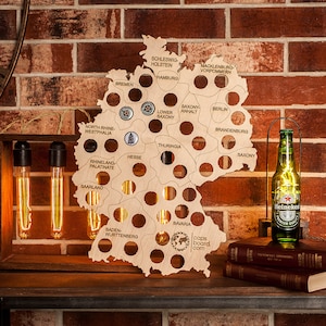 May include: Wooden map of Germany with cut-out circles, illuminated by lights. A Heineken beer bottle sits on a stack of books. A wooden frame with three Edison light bulbs is on the left. The background is a brick wall.