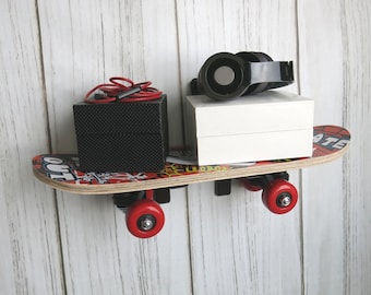 May include: A wall-mounted shelf shaped like a skateboard deck with red wheels. A black textured box with red wired earbuds sits on the left, and a white box with black over-ear headphones rests on the right side of the wooden skateboard surface.