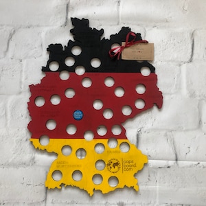 May include: A wooden map of Germany with bottle cap holes. The map is painted in the colors of the German flag: black, red, and yellow. The map is labeled with the names of the German states. The text "caps board.com" is printed in the lower right corner.