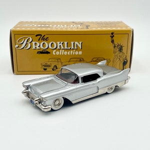 Könnte beinhalten: Ein silbernes 1959 Cadillac Coupe DeVille Modellauto aus der The Brooklyn Collection. Das Auto steht auf einer weißen Oberfläche mit einer Schachtel dahinter. Die Schachtel ist gelb mit schwarzem Text, der "The Brooklyn Collection" lautet.