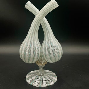 Puede incluir: Un florero de vidrio de doble cuello con dos cámaras bulbosas en forma de lágrima que se cruzan en la parte superior. El vidrio es transparente con rayas verticales blancas y azul claro y un patrón texturizado entrecruzado sobre una base.
