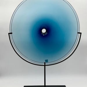 May include: A blue glass disc with a gradient of color from light blue to dark blue. The disc is mounted on a black metal stand with a round base.