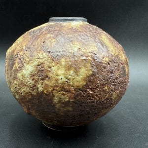 Könnte beinhalten: Eine runde, braune und gelbe Keramikvase mit einer strukturierten Oberfläche. Die Vase hat eine kleine Öffnung oben.