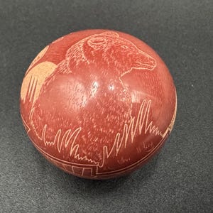 May include: A red clay pot with a carved design of a bear in a woodland setting.