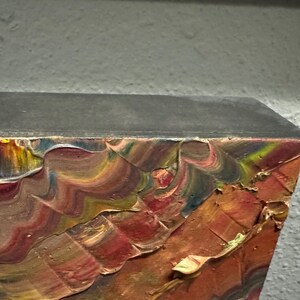 Puede incluir: Primer plano de una esquina de una pintura con pinceladas gruesas y texturizadas. Los colores incluyen tonos de rojo, naranja, amarillo, azul y verde. El borde superior es gris oscuro y el fondo es gris claro.