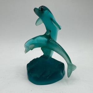 May include: A translucent teal glass sculpture featuring two dolphins in a dynamic leaping pose. One dolphin is positioned vertically while the other is arched horizontally, both mounted on a solid, textured teal glass base with smooth edges.