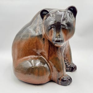 May include: A ceramic bear figurine featuring a glossy finish with blended tones of deep brown, black, and warm copper. The bear is depicted in a seated position with its head turned slightly to the side, showcasing smooth, rounded contours.