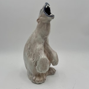 May include: A glazed ceramic figurine of a polar bear standing on its hind legs with its mouth wide open. The bear is colored in shades of off-white and light gray, with a dark blue interior inside the mouth and visible white teeth.