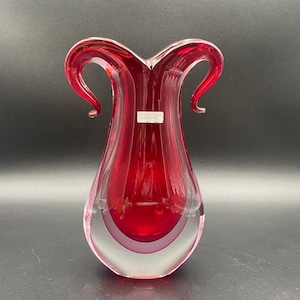May include: A red glass vase with a clear glass base and two curved handles. The vase has a label that reads "Evolution - Designed".