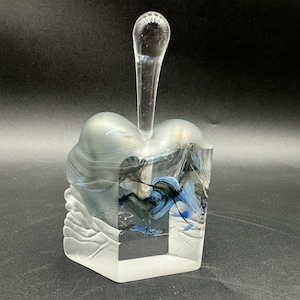 May include: A clear glass bottle stopper with a unique, abstract design featuring swirls of blue and black glass. The stopper has a tall, slender neck and a rounded top.