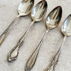 May include: Four antique silver spoons with ornate handle designs. The spoons have a reflective surface and are arranged diagonally. The handles feature floral and scroll patterns, and some have engraved initials. The spoons are a vintage tableware set.