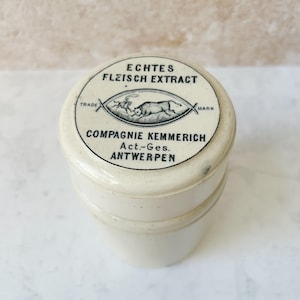 May include: An antique, off-white jar with a screw-on lid. The lid features black text and a graphic of a bull and other animals. The text reads "ECHTES FLEISCH EXTRACT" and "COMPAGNIE KEMMERICH, Act.-Ges., ANTWERPEN."