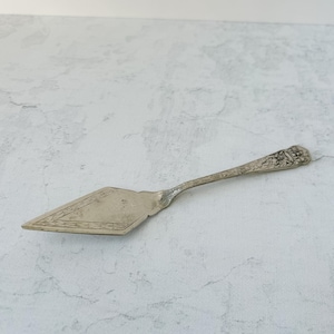 May include: A vintage silver pie server with a diamond-shaped blade and a decorative handle. The blade features an etched border design. The handle has an ornate floral and scroll pattern. The server is made of a shiny, reflective metal.