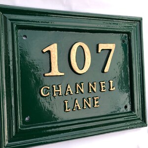 May include: A rectangular, dark green house number sign with a gold border. The number "107" and the words "CHANNEL LANE" are in gold lettering. The sign has a glossy finish and is mounted on a white background.