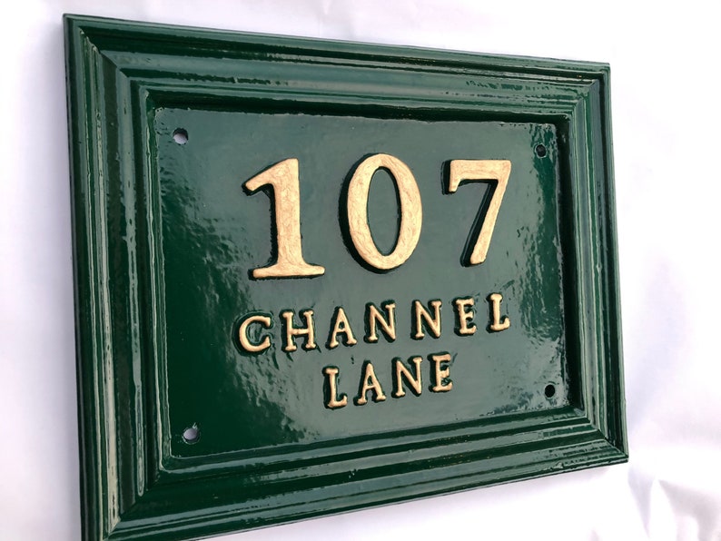 May include: A rectangular, dark green house number sign with a gold border. The number "107" and the words "CHANNEL LANE" are in gold lettering. The sign has a glossy finish and is mounted on a white background.