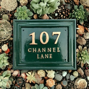 May include: A rectangular, dark green address plaque with the numbers "107" and the words "CHANNEL LANE" in gold lettering. The plaque is surrounded by a border and set among small rocks and succulents.