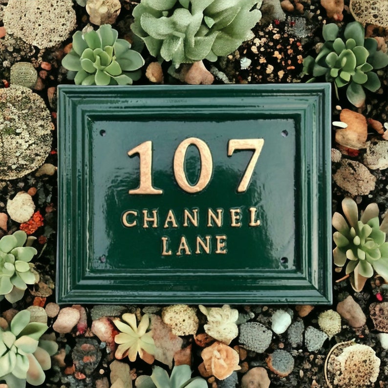 May include: A rectangular, dark green address plaque with the numbers "107" and the words "CHANNEL LANE" in gold lettering. The plaque is surrounded by a border and set among small rocks and succulents.