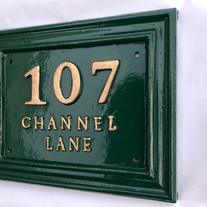 May include: A rectangular, dark green house number sign with gold-colored raised lettering. The sign reads "107 CHANNEL LANE". The sign has a glossy finish and a decorative border.