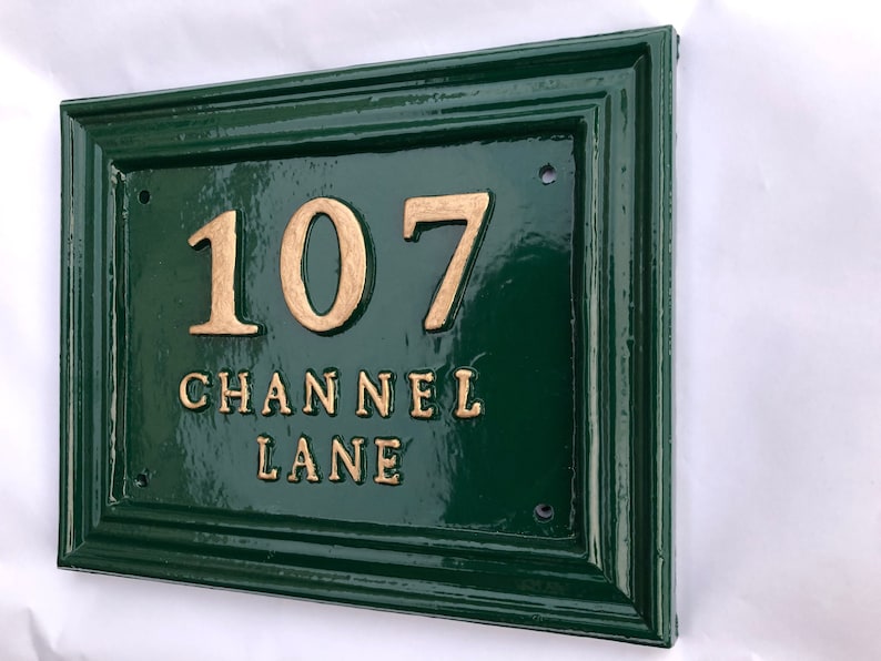 May include: A rectangular, dark green house number sign with gold-colored raised lettering. The sign reads "107 CHANNEL LANE". The sign has a glossy finish and a decorative border.