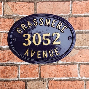 May include: A blue oval-shaped house number sign with gold lettering. The sign reads "Grassmere 3052 Avenue".