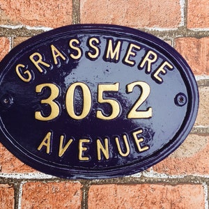 May include: Oval-shaped house number sign with a blue background and gold lettering. The sign reads "GRASSMERE 3052 AVENUE".