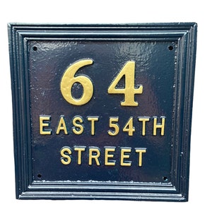 以下が含まれることがあります： 金色の文字が入った長方形の濃紺の住所プレート。「64 EAST 54TH STREET」と書かれています。数字と文字は浮き上がっています。プレートには装飾的なボーダーがあります。