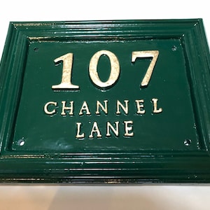 May include: Green rectangular address plaque with gold lettering. The plaque reads "107 CHANNEL LANE". The frame is a darker green. The numbers and letters are raised.