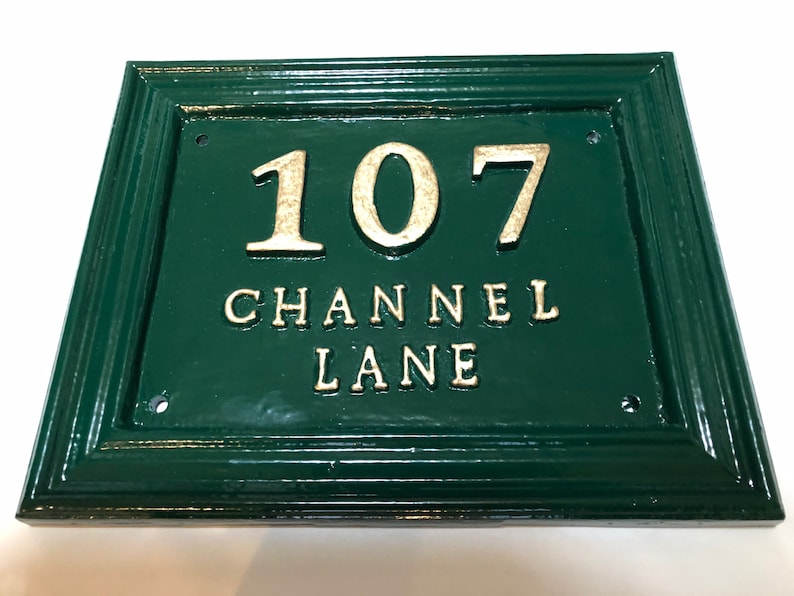 May include: Green rectangular address plaque with gold lettering. The plaque reads "107 CHANNEL LANE". The frame is a darker green. The numbers and letters are raised.